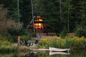 Une cabane dotée d’une terrasse en bois, entourée d’une forêt et bordée par un plan d’eau.