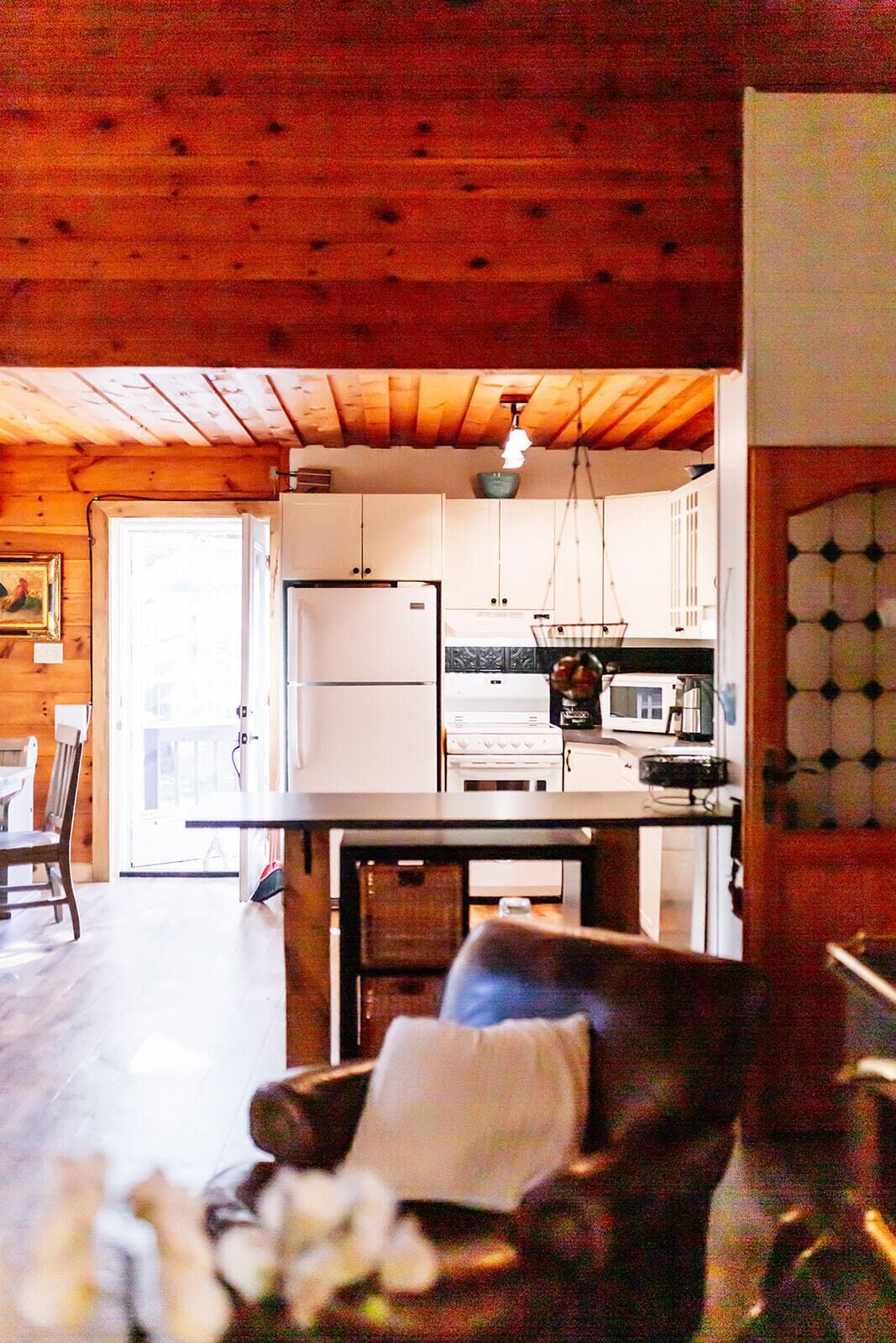 Un interior de cabaña acogedor, con techo de madera, zona de comedor con mesa y sillas, cocina con estufa y refrigerador, y sala de estar con sillón y un jarrón de flores.