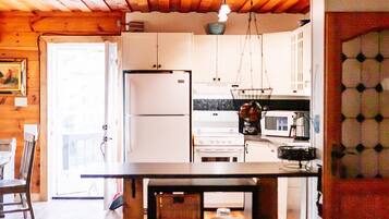 Un interior de cabaña acogedor, con techo de madera, zona de comedor con mesa y sillas, cocina con estufa y refrigerador, y sala de estar con sillón y un jarrón de flores.