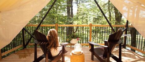 A tented outdoor area with two Adirondack chairs and a table, surrounded by a wooden deck and a view of a forest.