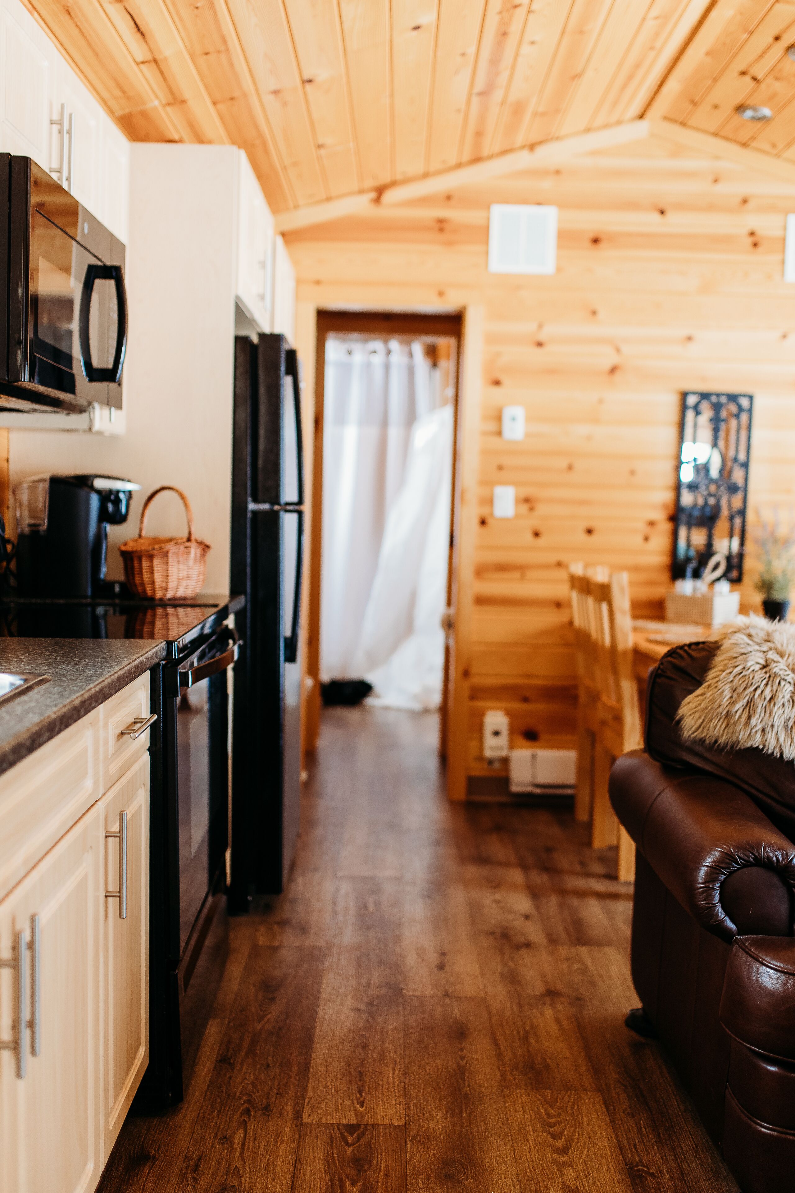 Interior de madera con cocina, que incluye armarios, microondas y refrigerador. Hay una mesa de comedor con sillas y un sofá de cuero marrón.