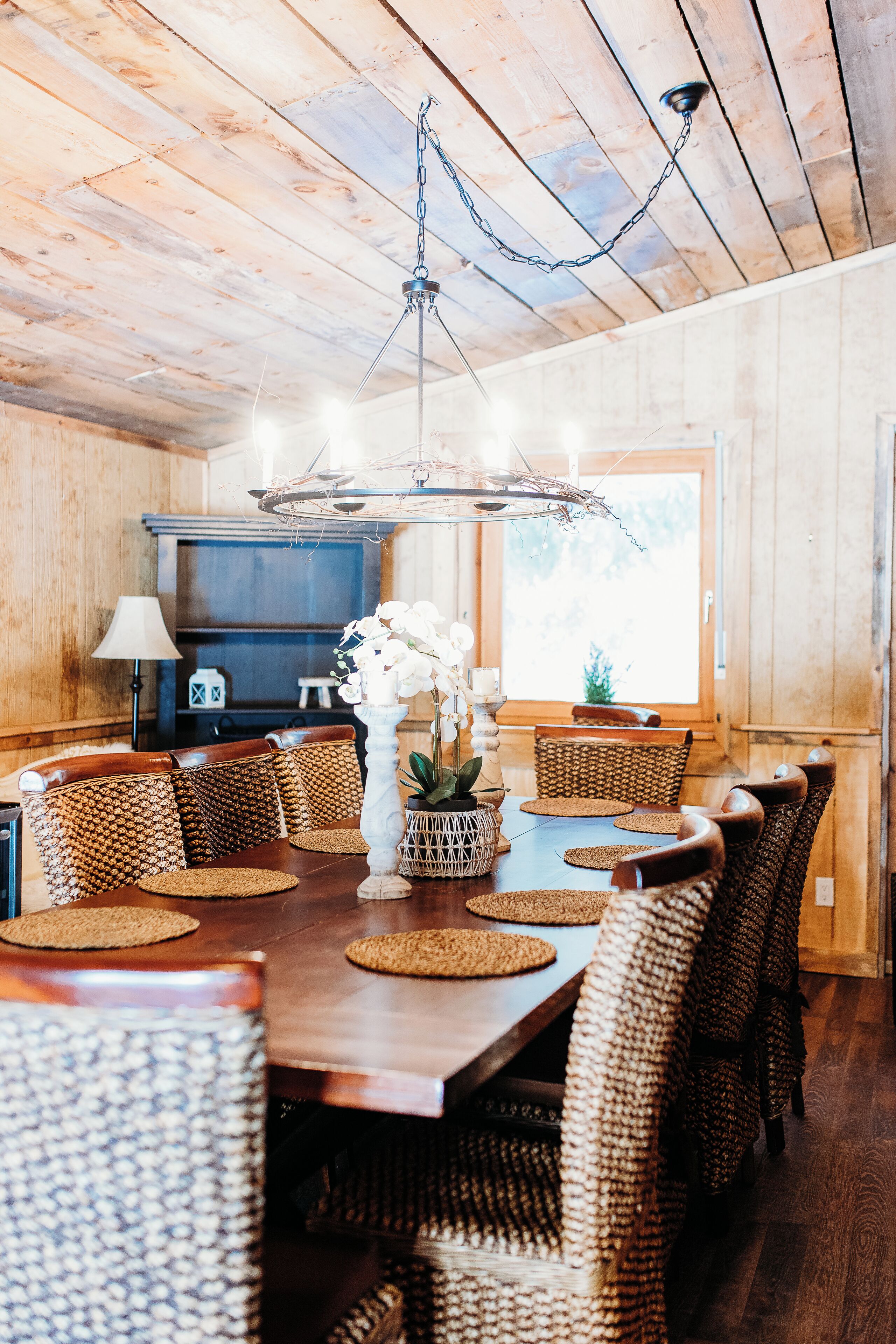 Une salle à manger aux murs lambrés, avec une grande table, des chaises en osier et un luminaire suspendu.
