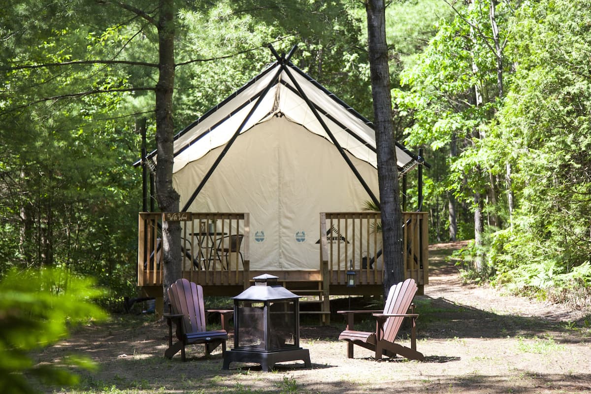 Ein Waldcampingplatz mit einem großen Zelt, einer Holzterrasse und mehreren Adirondack-Stühlen.