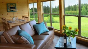 Een gezellig interieur van een chalet met een leren bank, een eettafel en grote ramen met uitzicht op een groen landschap.