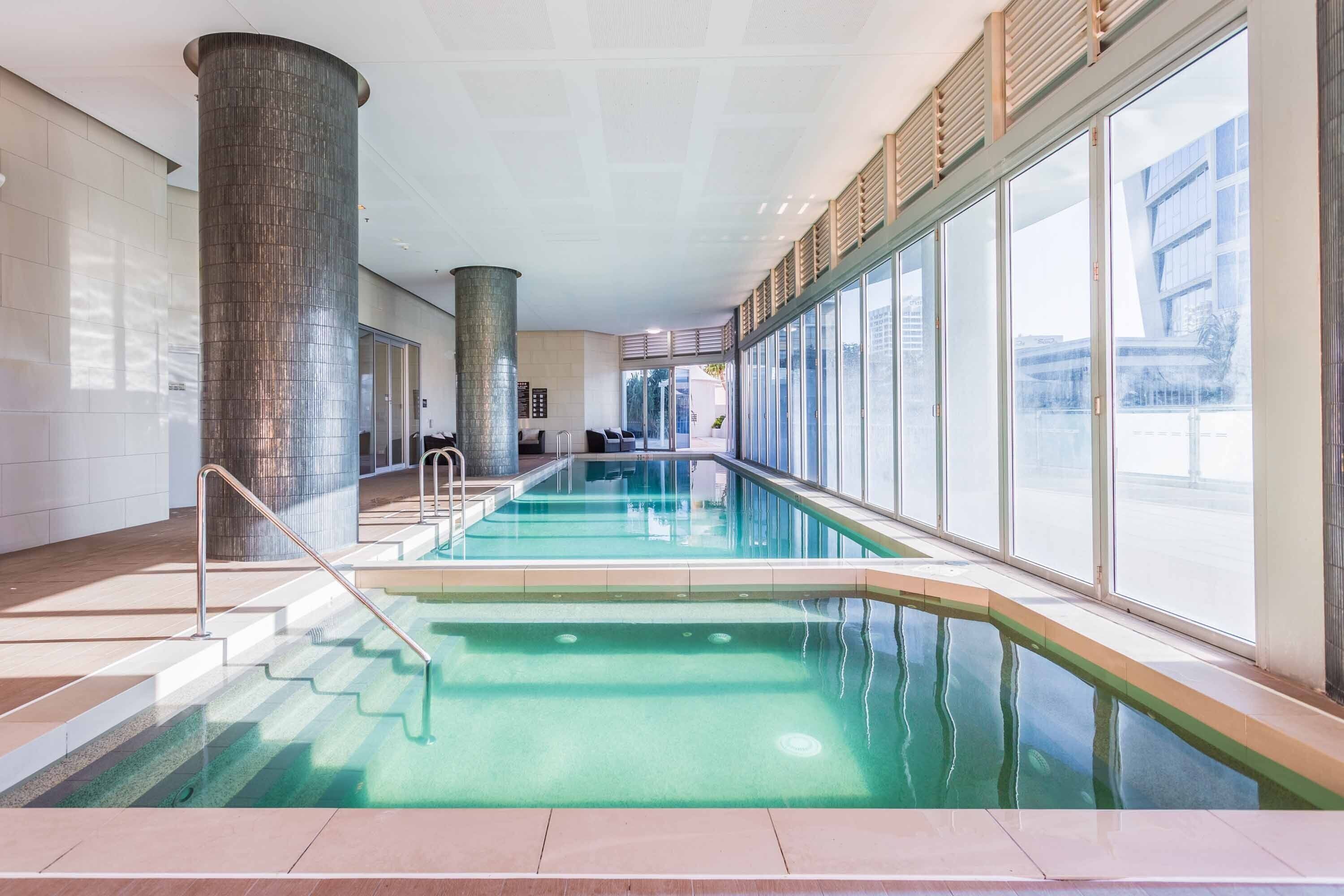 En indendørs swimmingpool med glasvægge, en central søjle og en trappe ned til poolen.