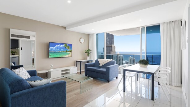 A modern living room with a sofa, coffee table, and a dining area with a view of the city and ocean. - H Residences Ocean View (Surfers Paradise)