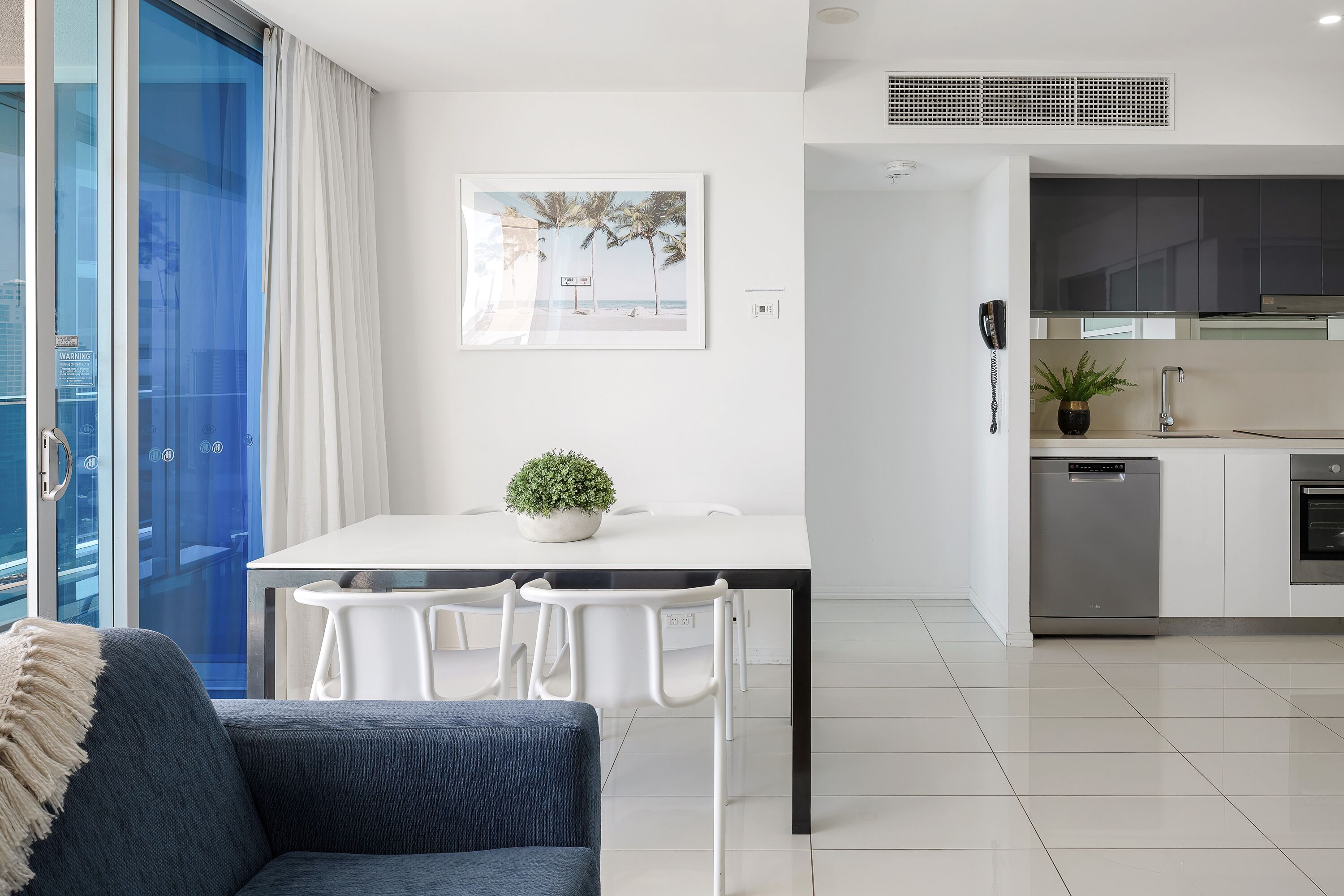 A modern living room with a sofa, a dining table with chairs, a kitchen area with cabinets and appliances, and a large window with curtains.