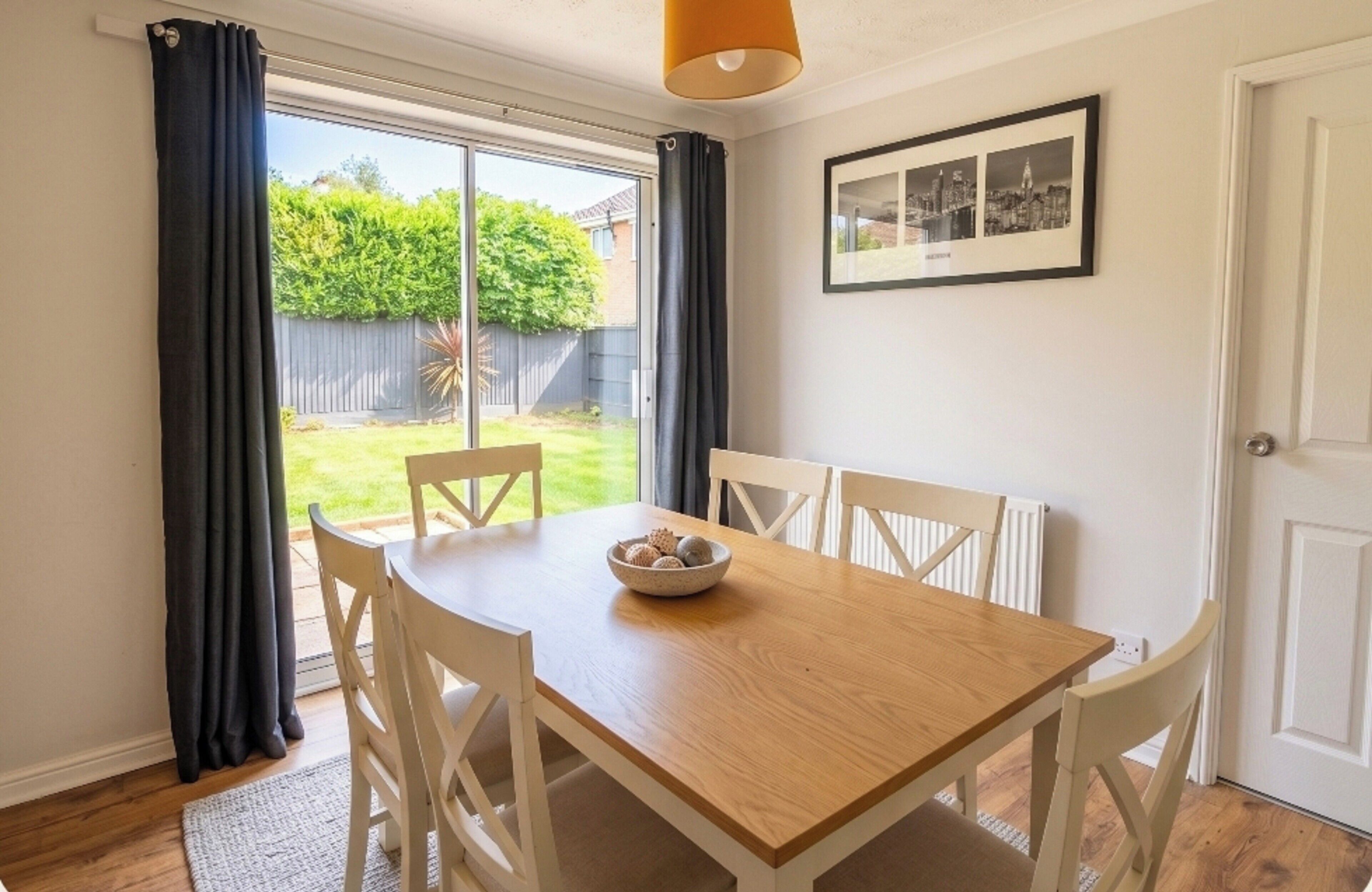 Uma sala de jantar com uma mesa de madeira, cadeiras brancas, uma tigela de petiscos e uma fotografia de paisagem urbana na parede.
