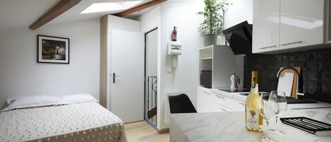 A bedroom with a bed, a small kitchenette with a wine bottle and glasses, and a bathroom with a shower.
