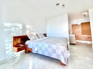 A modern bedroom with a bed, a wooden bench, a brick accent wall, and a bathroom with a sink and mirror. - Inviting 2-Bed Apartment in Cofresi (Cofresi, Puerto Plata)