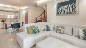 Una sala de estar moderna con un sofá blanco, una mesa de comedor con sillas, una cocina al fondo y una escalera que sube a otro nivel.