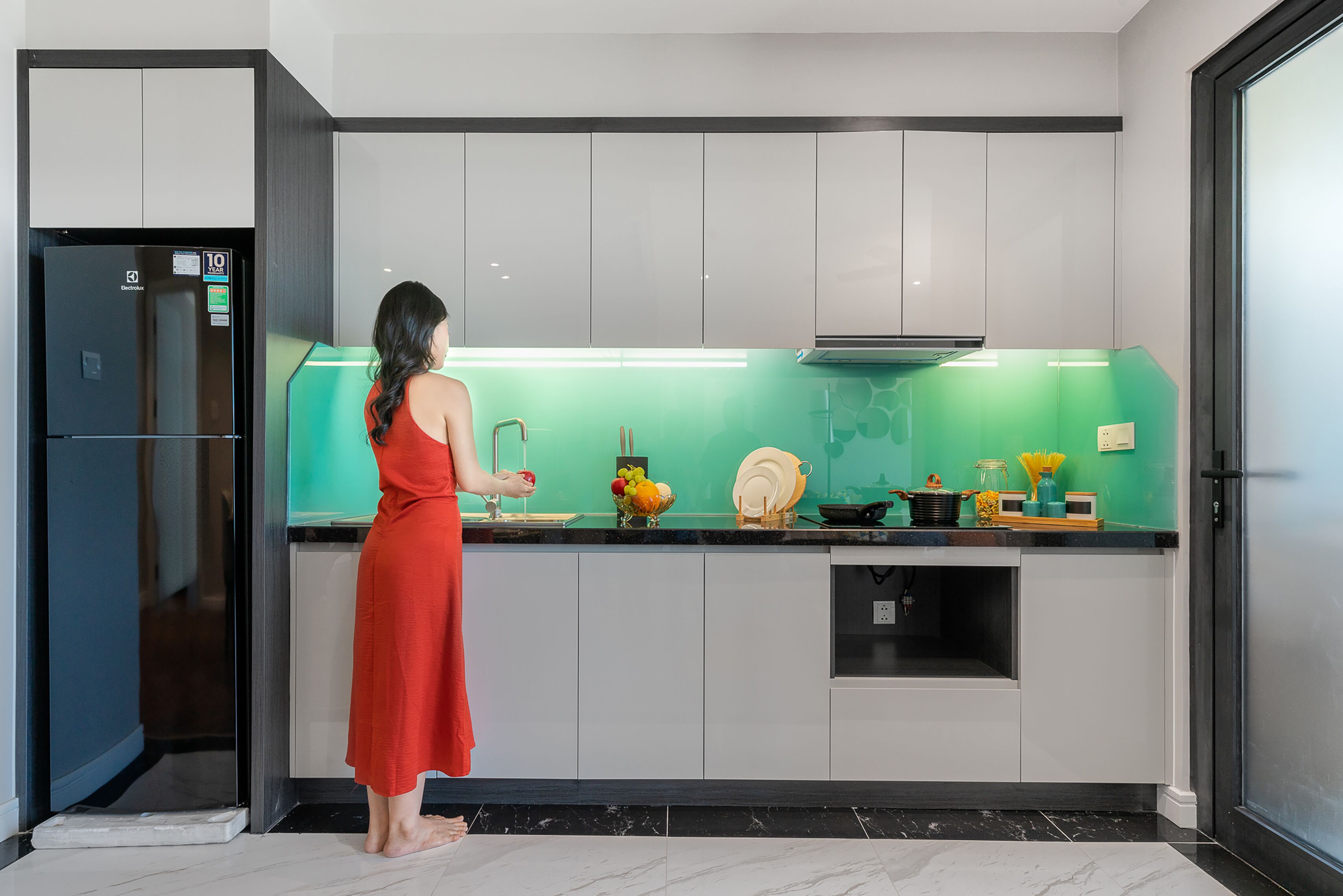 A modern kitchen with a woman in a red dress, a black refrigerator, and a green backsplash.
