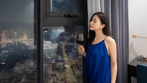 Une personne se tient près d’une fenêtre, d’où l’on peut admirer un panorama sur la ville, et elle tient un verre de vin à la main.