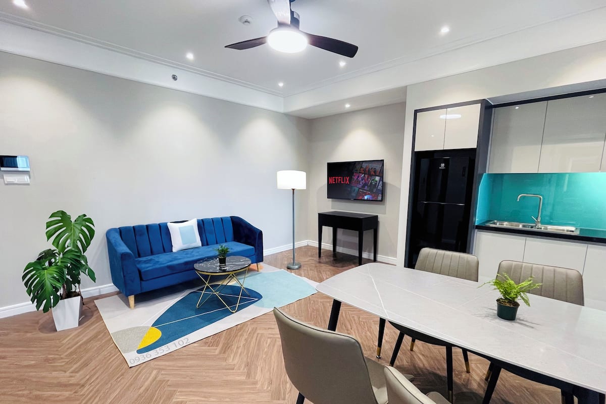 A modern living room with a blue sofa, a dining table with chairs, a TV, and a kitchen area with a sink.