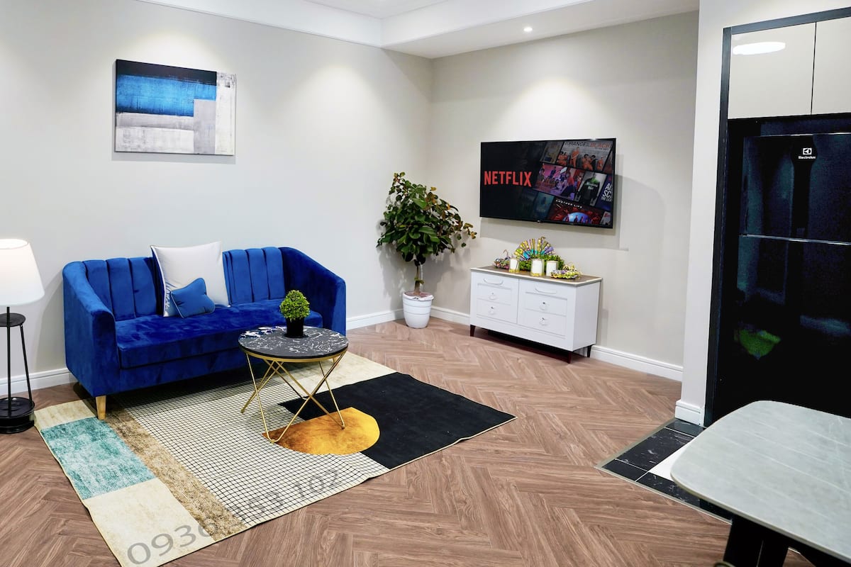 A modern living room with a blue sofa, a small round table, a potted plant, a TV displaying Netflix, and a herringbone-patterned floor.