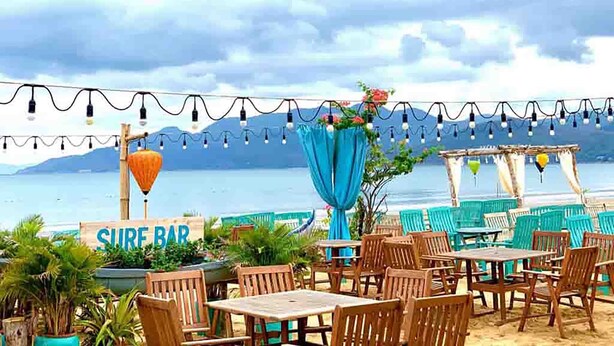 Un espace extérieur aménagé avec des tables et des chaises en bois, une enseigne indiquant la présence d’un bar à thème surf, et une vue sur la mer.