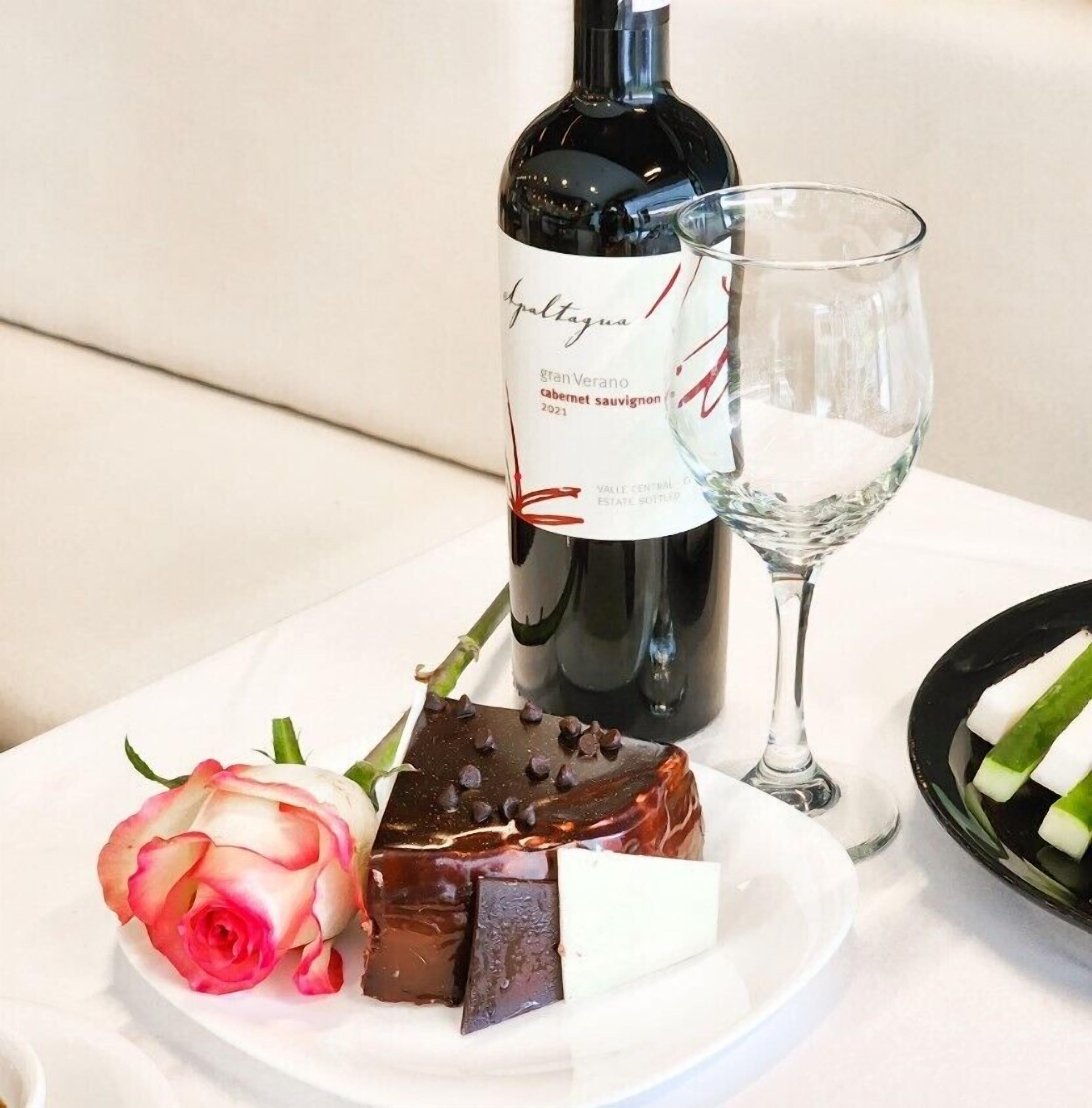 A bottle of wine, a glass, and a plate with chocolate and a rose.