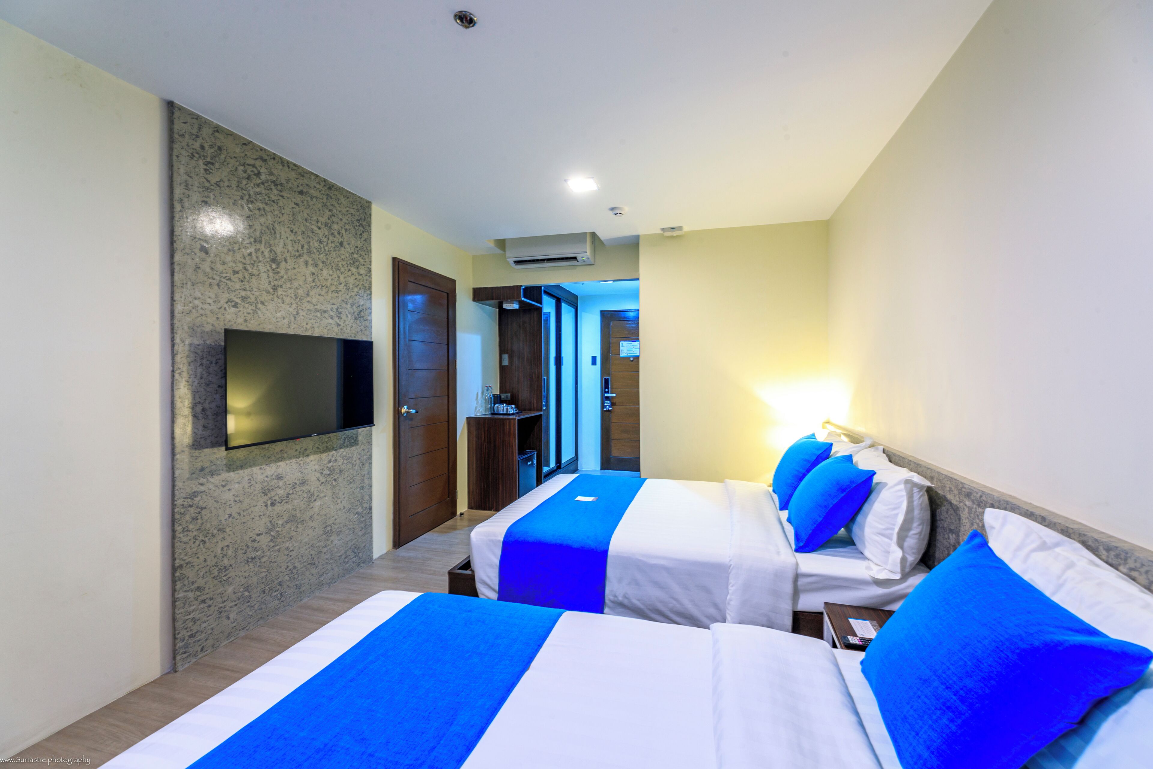 A hotel room with two beds, a flat-screen TV mounted on the wall, and a door leading to a hallway.