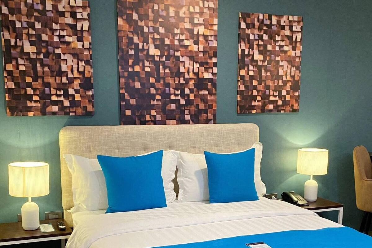 A hotel room with a bed, two bedside tables, a lamp, a chair, and three wall art pieces.