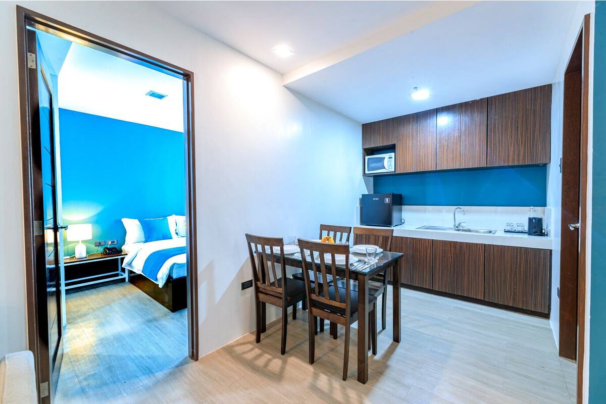 A compact hotel room with a bed, bedside table, and a small kitchenette.