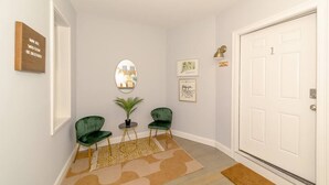 Un couloir avec deux chaises vertes, une petite table, un miroir et des œuvres d’art encadrées accrochées aux murs.