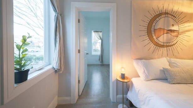 Une chambre à coucher comprenant un lit, une table de chevet avec une lampe, une œuvre d’art accrochée au mur et une fenêtre avec des rideaux.