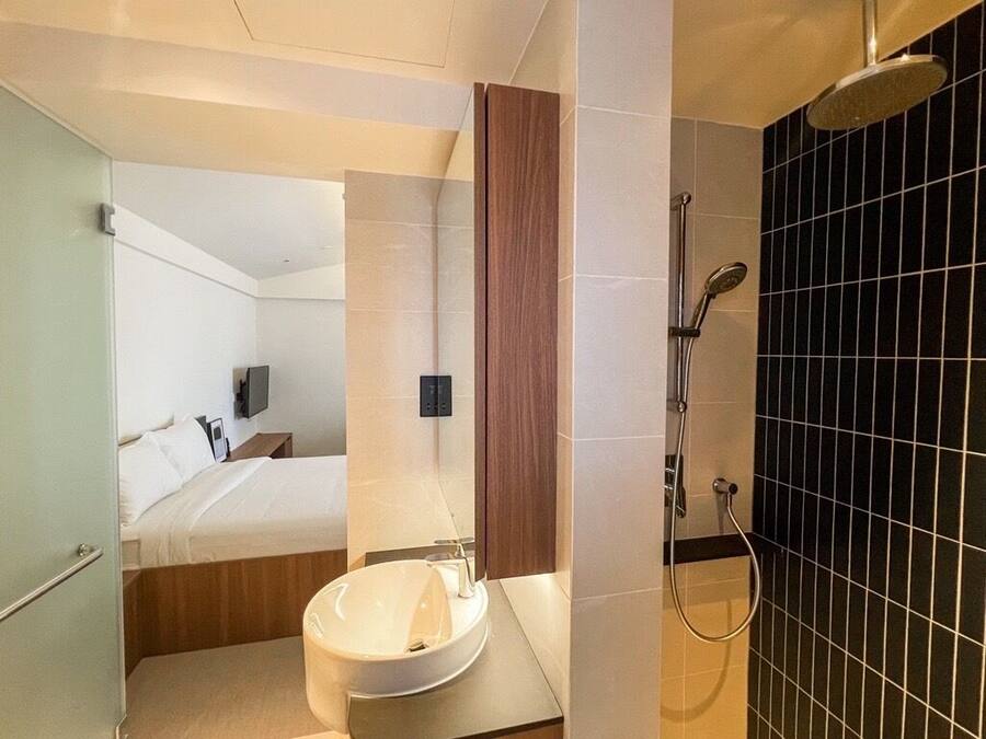 Un baño moderno con un lavamanos grande, ducha y una cama a la vista en el fondo.