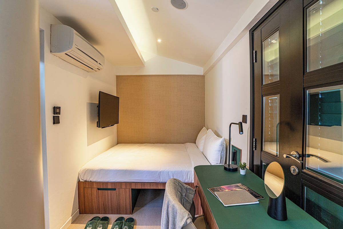 A compact hotel room with a single bed, a desk with a chair, a small table, a mirror, a TV mounted on the wall, and an air conditioning unit.