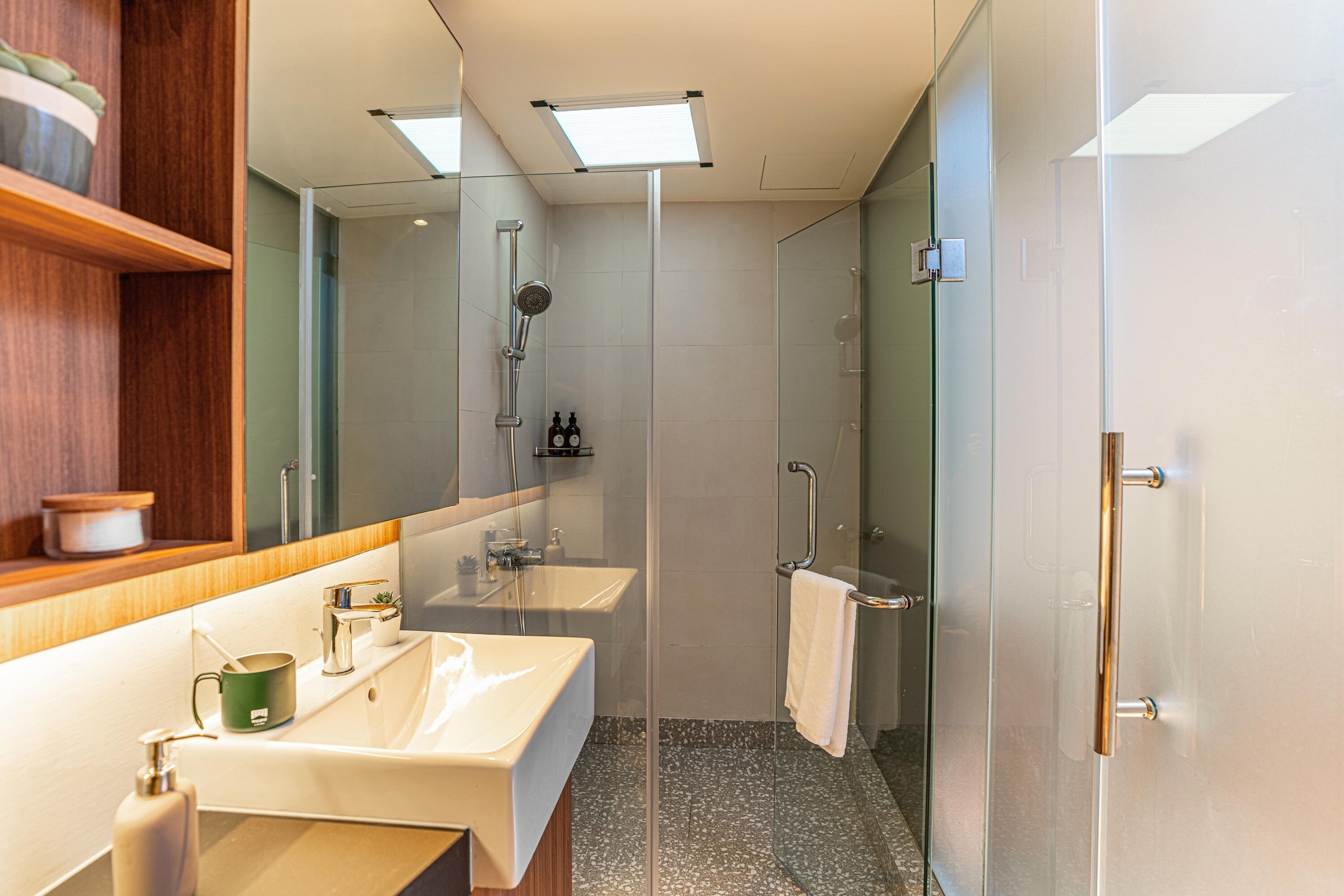A modern bathroom with two sinks, a glass shower enclosure, and a wooden shelf with toiletries.