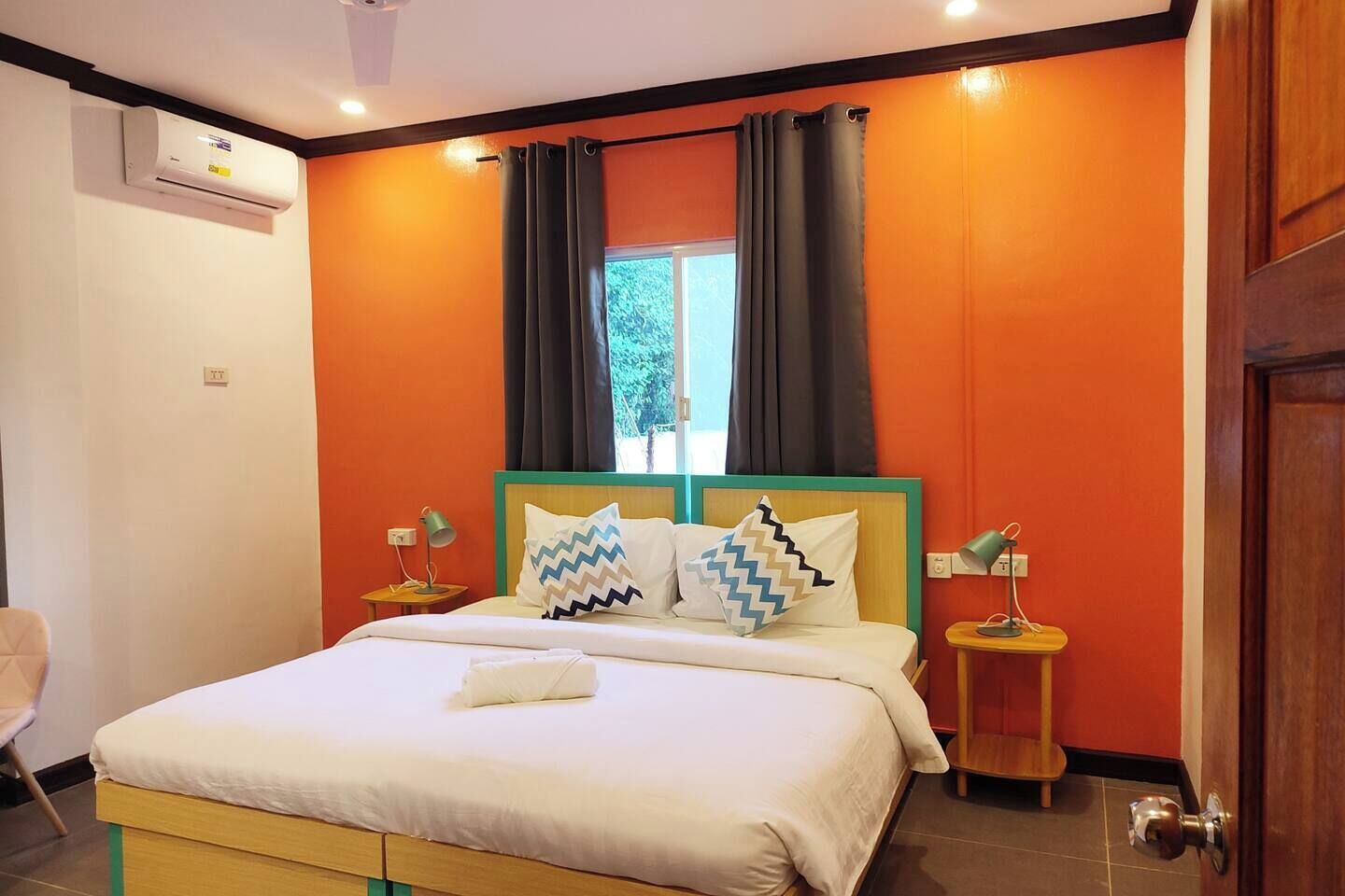 A hotel room with a bed, two bedside tables, an air conditioning unit, and a window with curtains.