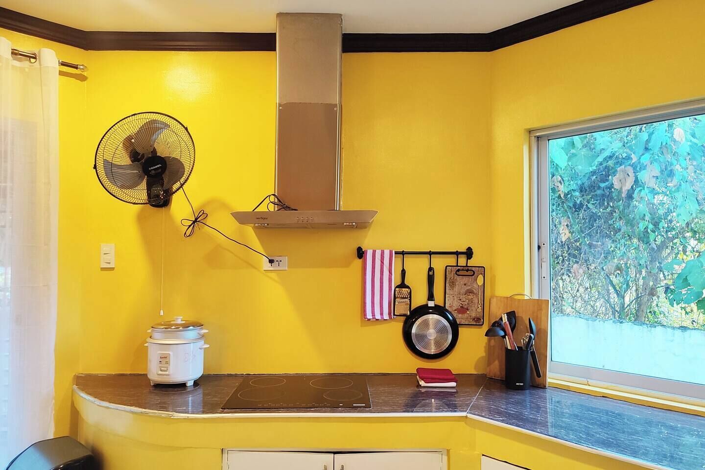 A kitchen with a yellow wall, a fan, a stove, and a window with a view of trees.
