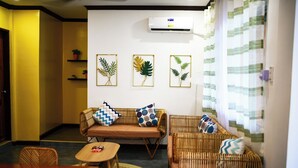 Una sala de estar con un sofá, dos sillones, una mesa de centro y una mesita con un libro. Hay tres obras de arte con hojas enmarcadas en la pared y un equipo de aire acondicionado sobre el sofá.