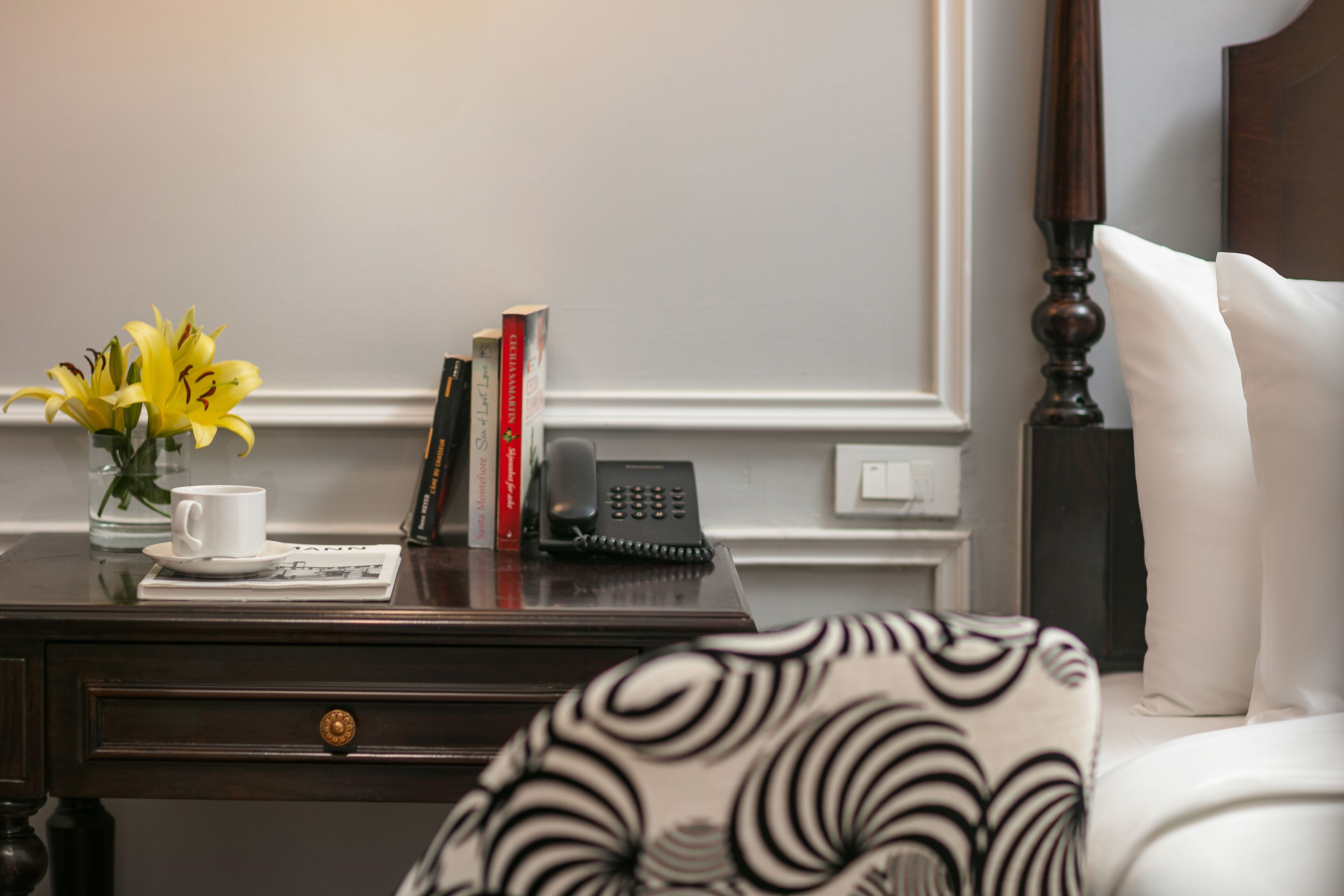 A neatly arranged bedroom with a bedside table holding a vase of yellow flowers, a cup, a book, and a telephone.