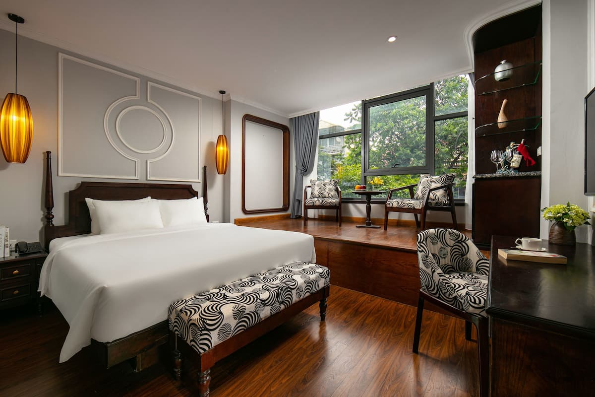 Une chambre d’hôtel avec un grand lit, un banc en bois, un bureau avec une tasse et un vase, et une vue sur l’extérieur par une grande fenêtre.