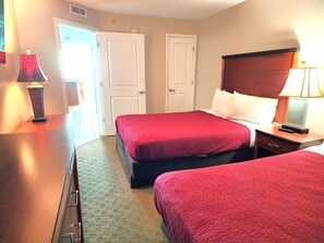 Habitación de hotel con dos camas, un aparador de madera, una mesita de noche con lámpara y una puerta que da a otra habitación.