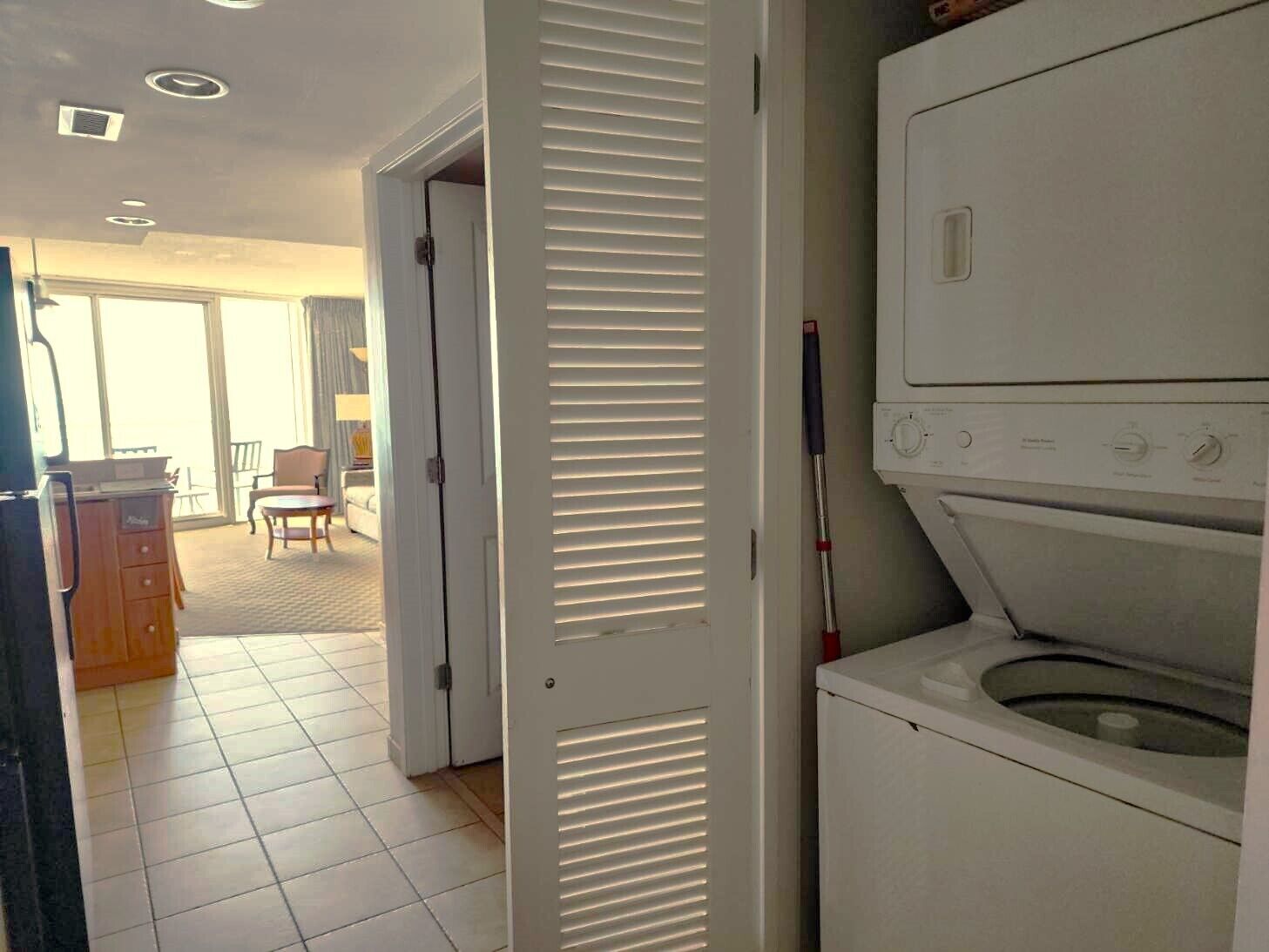 Een wasserette met een wasmachine en droger, een deur naar een eetruimte, en een raam met jaloezieën.