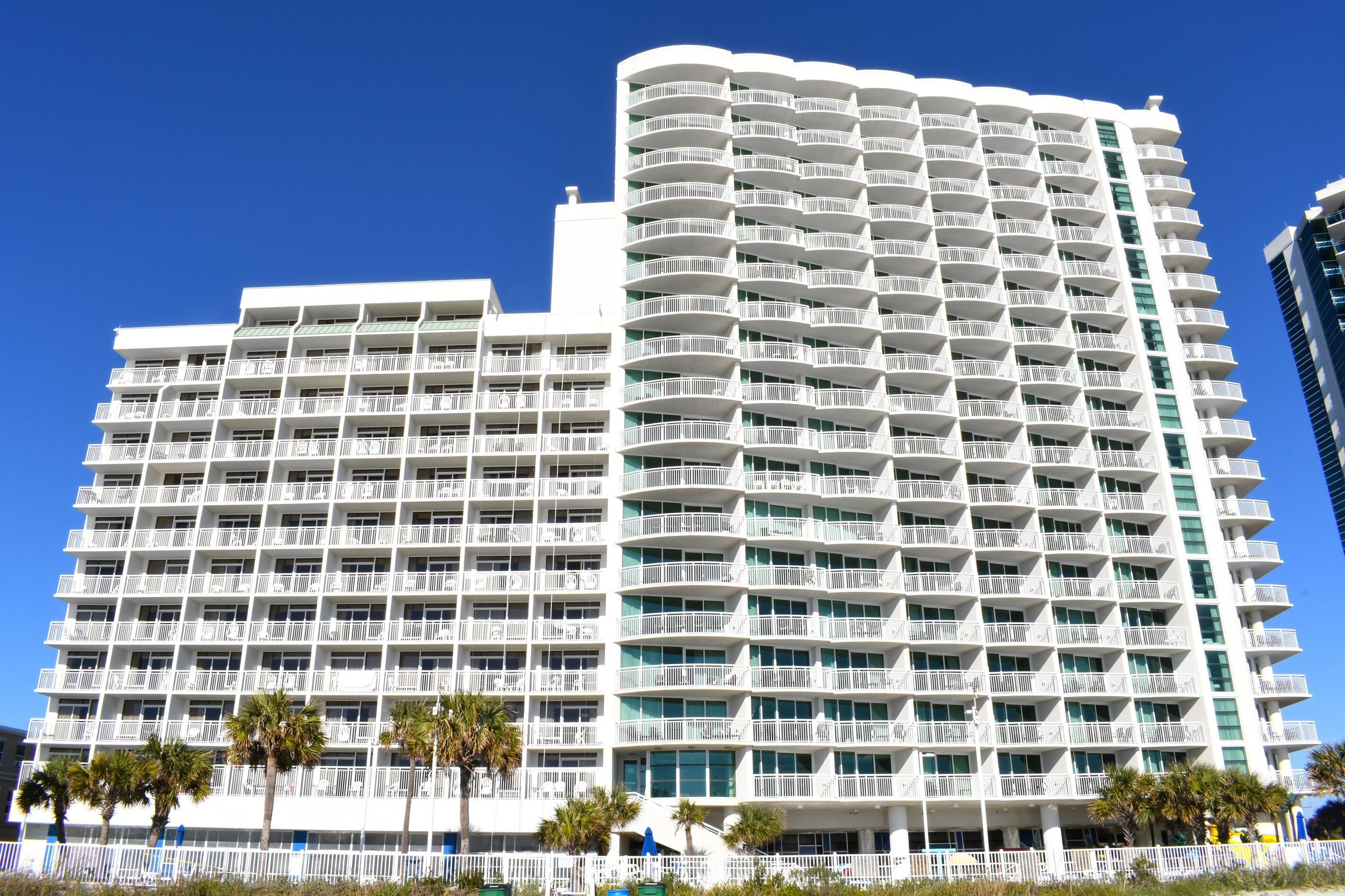 A multi-story hotel with balconies, palm trees in front, and a clear blue sky.
