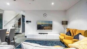 A modern living room with a yellow sofa, a flat-screen TV mounted on the wall, and a staircase leading to another level. - Wow Large 4 Bedrooms 4 bathroom with Private pool, 4 min walk to beach (Surfers Paradise)