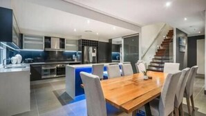 A modern kitchen with a central island, a dining area with a wooden table and chairs, and a staircase leading to an upper level. - Wow Large 4 Bedrooms 4 bathroom with Private pool, 4 min walk to beach (Surfers Paradise)