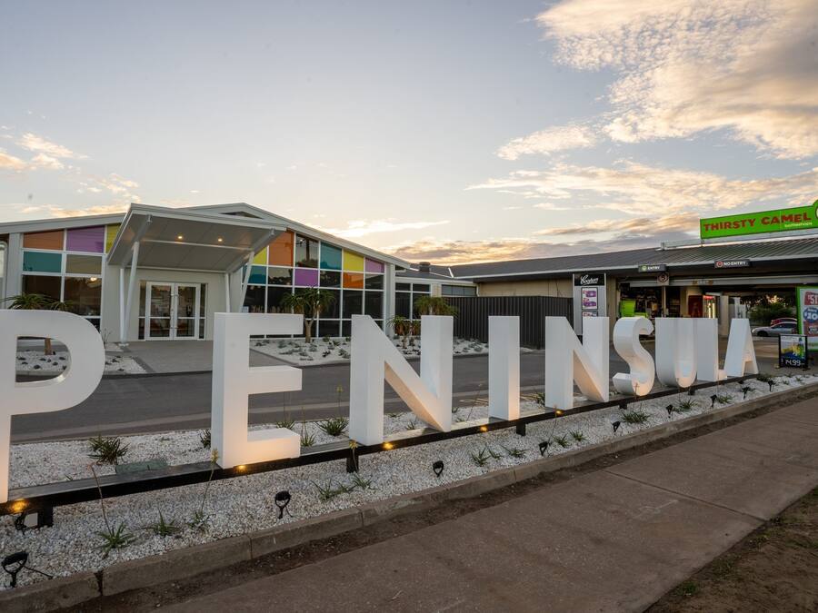 Ein modernes Gebäude mit großen weißen Buchstaben, die "PENINSULA" zeigen, und ein Schild für die Bar/das Restaurant "Thirsty Camel" im Hintergrund.