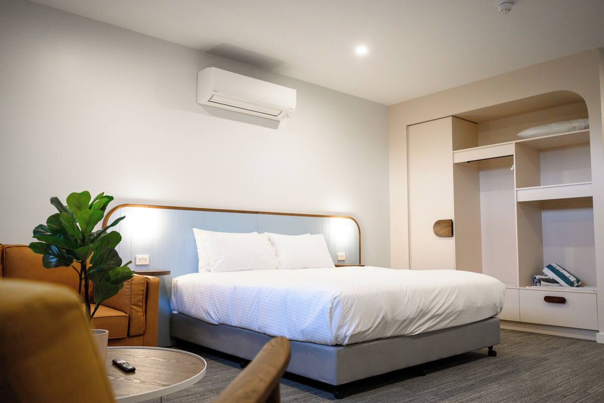 A hotel room with a bed, a bedside table, a chair, a plant, and a wardrobe.