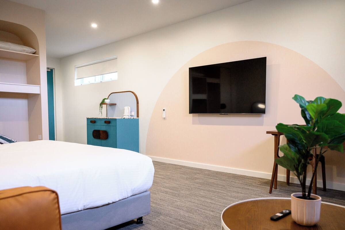 A hotel room with a bed, a TV mounted on the wall, a small table with a plant, and a blue cabinet.