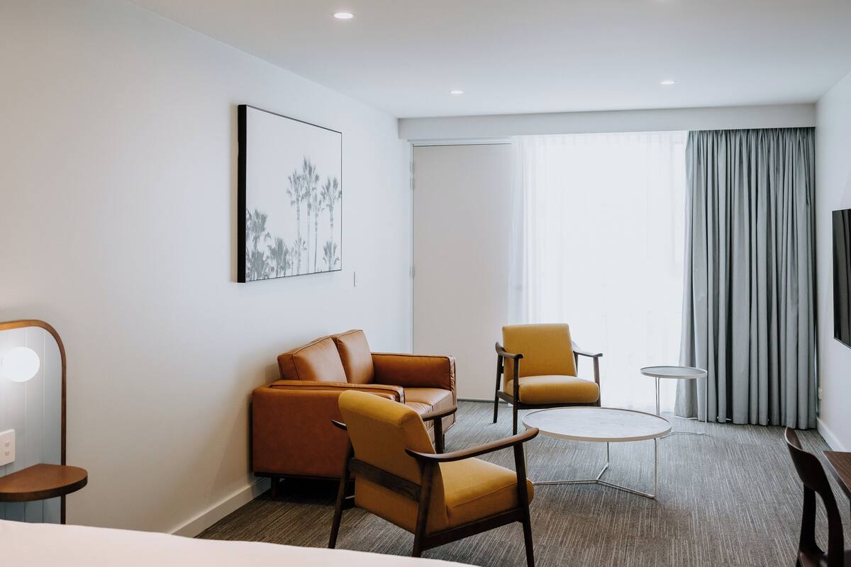 A modern hotel room with a large bed, a sofa, two armchairs, a small table, and a painting on the wall.