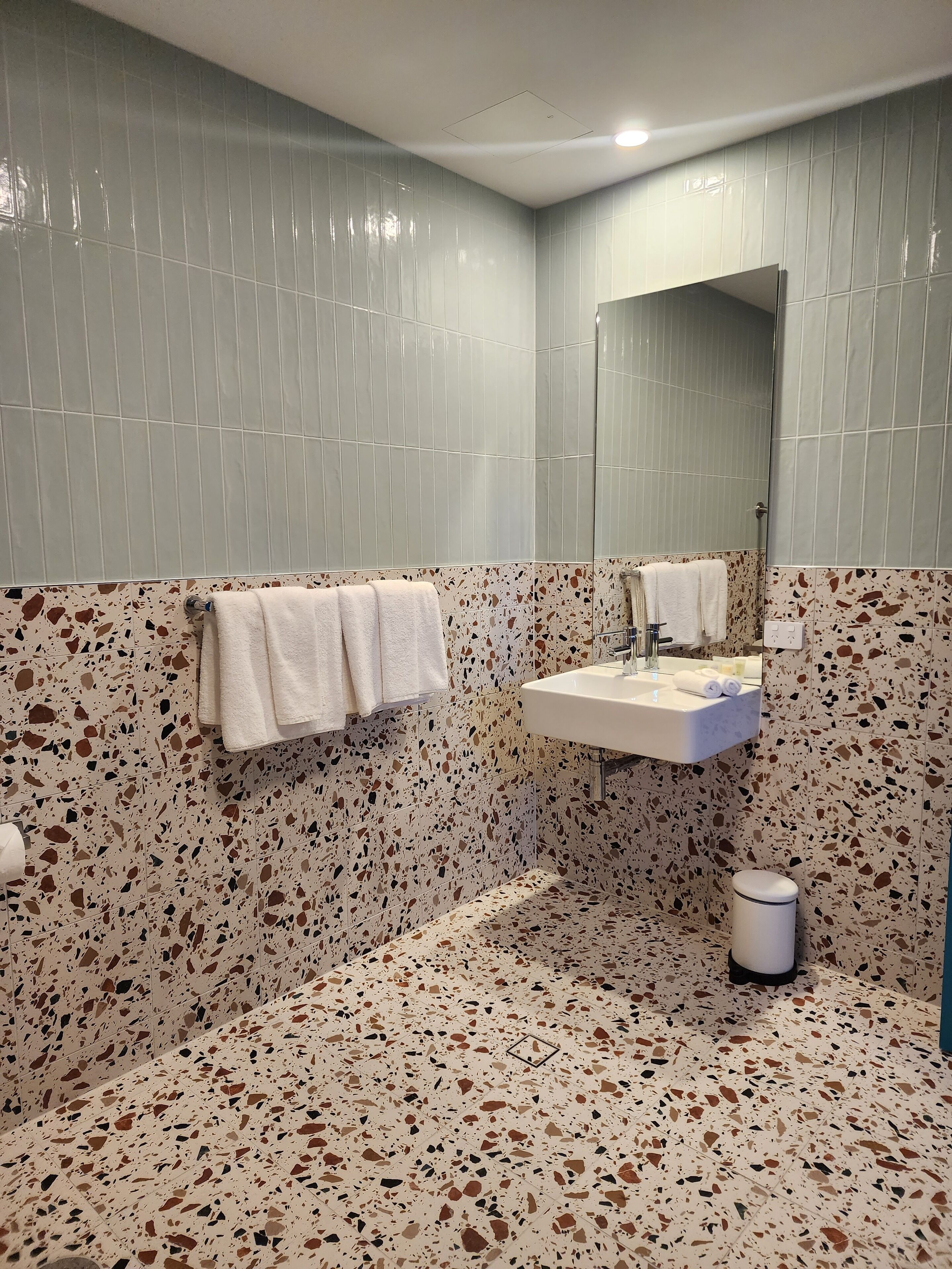 Een badkamer met een terrazzovloer, een wastafel met spiegel en een handdoekrek.