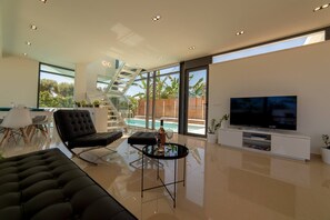 A modern living room with a large TV, a glass coffee table, a black sofa, and a staircase leading to a balcony.