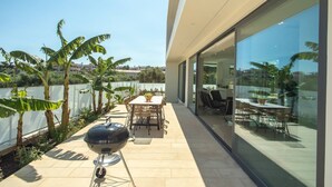 Un patio moderno con una parrilla, mesa de comedor y sillas, rodeado de paredes de vidrio y plantas tropicales.