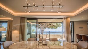 A modern dining area with a marble table and six chairs, large windows offering a view of the sea, and a chandelier with spherical glass shades.