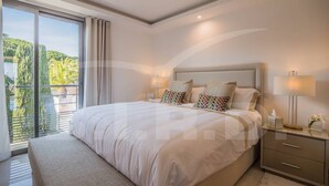 A modern bedroom with a large bed, a nightstand, and a view of greenery through the window.