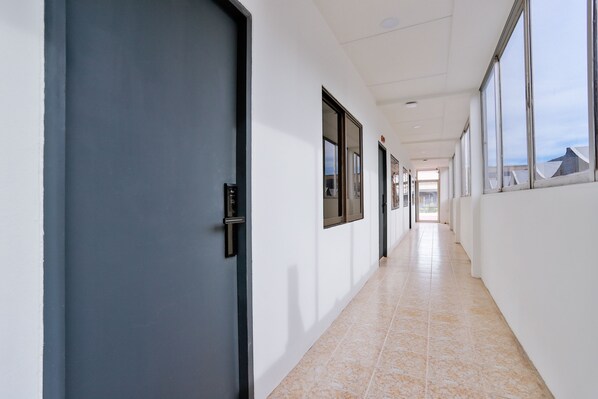 A long, narrow hallway with a blue door on the left, large windows on the right, and a tiled floor.