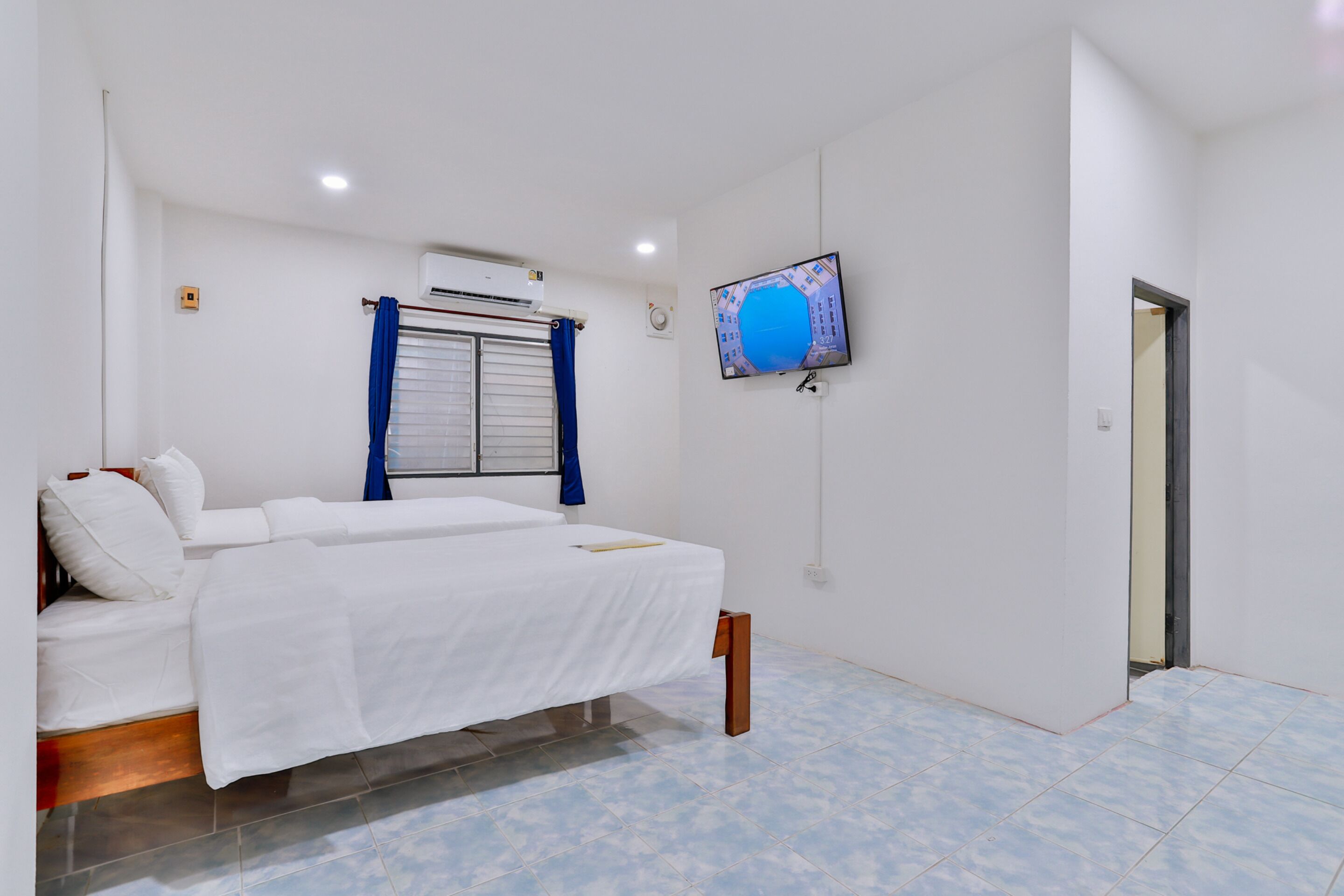 A bedroom with a bed, a TV mounted on the wall, an air conditioning unit, and a window with blue curtains.